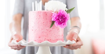 A tall pink cake with candles on the top and a small floral arrangement on the side sits on a curved cake tray.