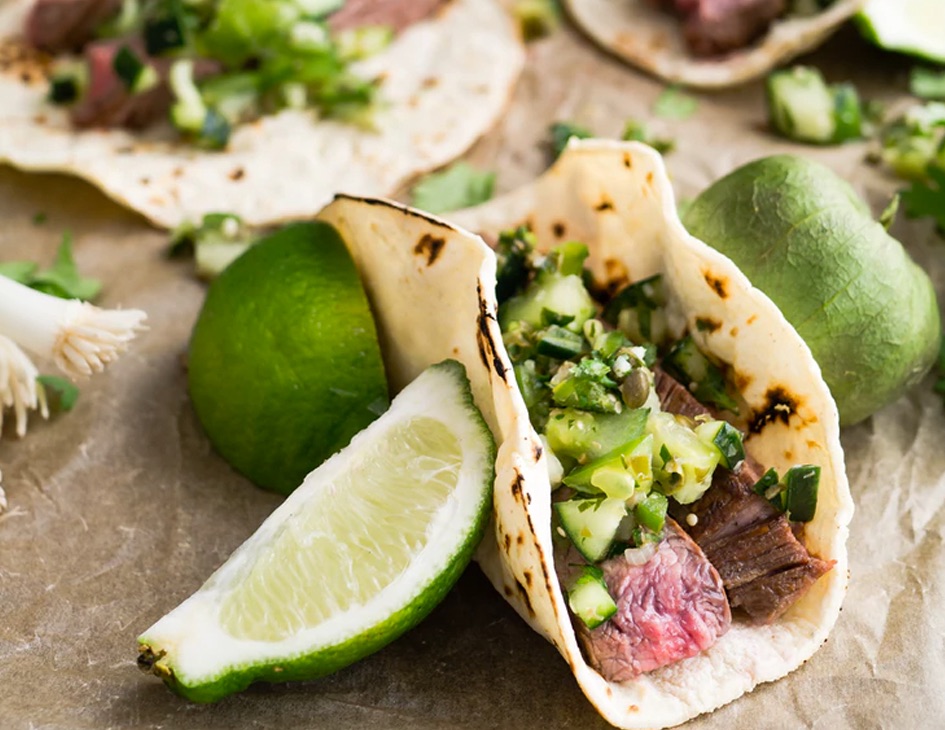 A taco filled with cucumber, cilantro, and rare beef is propped up by lime slices and a tomatillo.