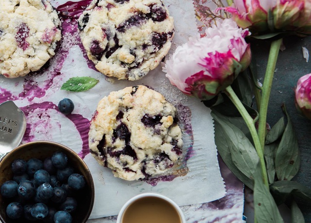 Some blueberry cookies lie on parchment paper next to a bowl of blueberries and a couple of flowers.